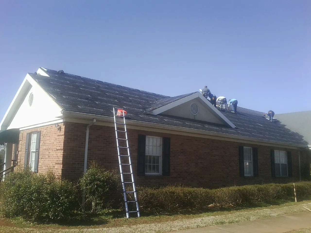 Crew tearing shingles off a brick ranch roof with a ladder set — Metal Roof Repair work by our crew in Riva