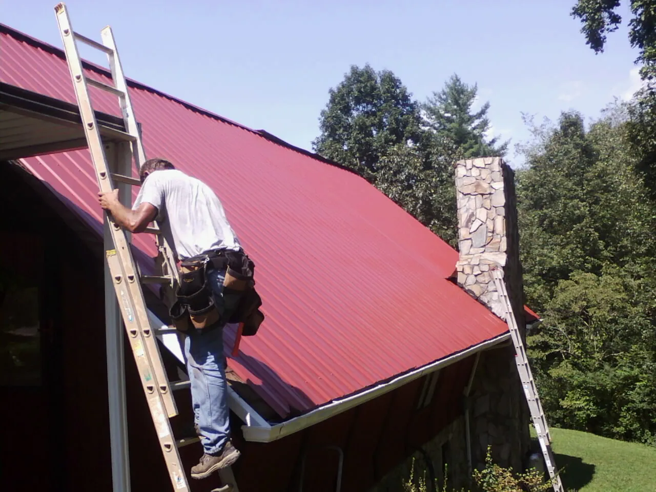 Roofer on a ladder beside a red metal roof and stone chimney — Soffit & Fascia Repair by Roofing Brothers in Riva